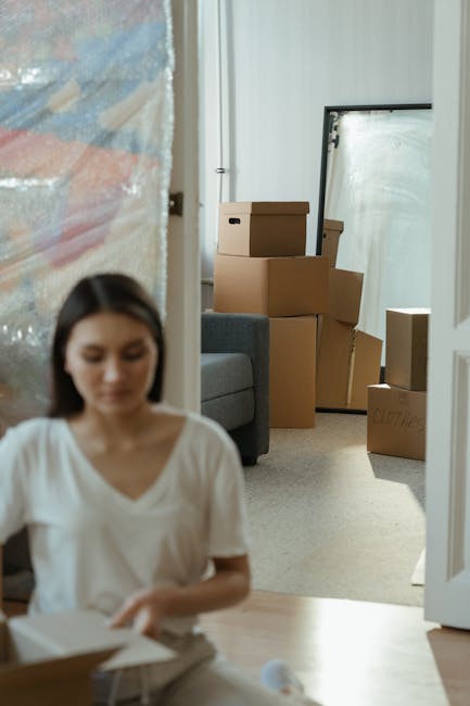 A person lying on the floor surrounded by multiple cardboard boxes of various sizes, some sealed with packing tape and others partially open, inside a well-lit room with large windows and a radiator b