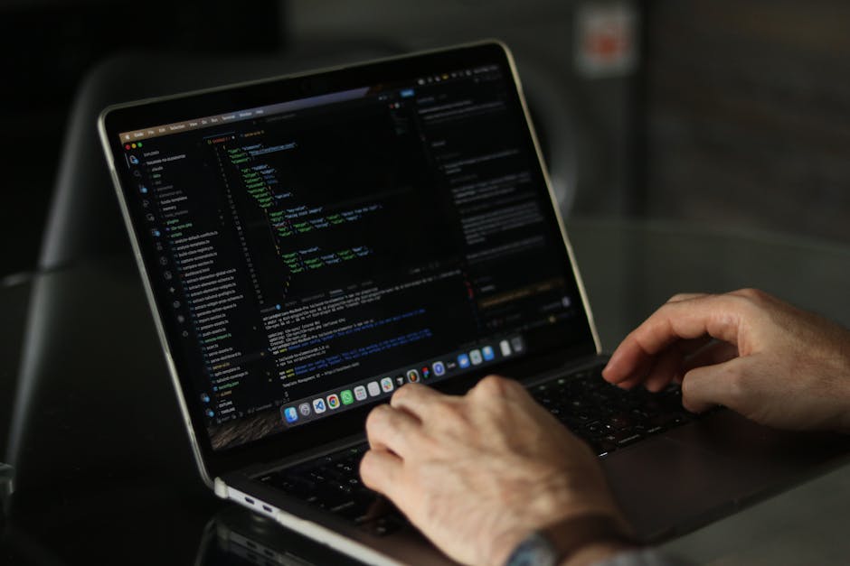 Close-up of a person's hands operating a laptop on a dark table, displaying the screen with visible lines of code and text, indicating a coding or programming session. The environment appears to be in
